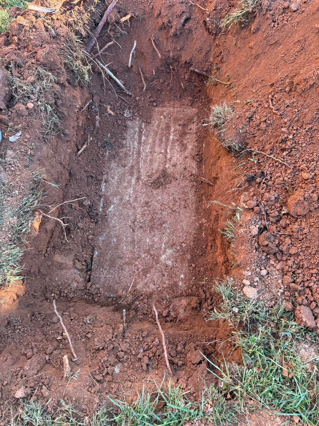 A rectangular, dirt-covered concrete slab is partially exposed within an open, rectangular trench in reddish-brown soil.