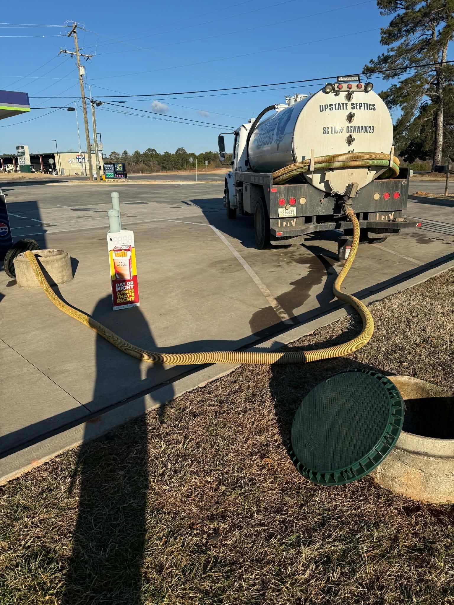 A tanker truck is parked at a service station, pumping fluid through a hose from an open underground access point.