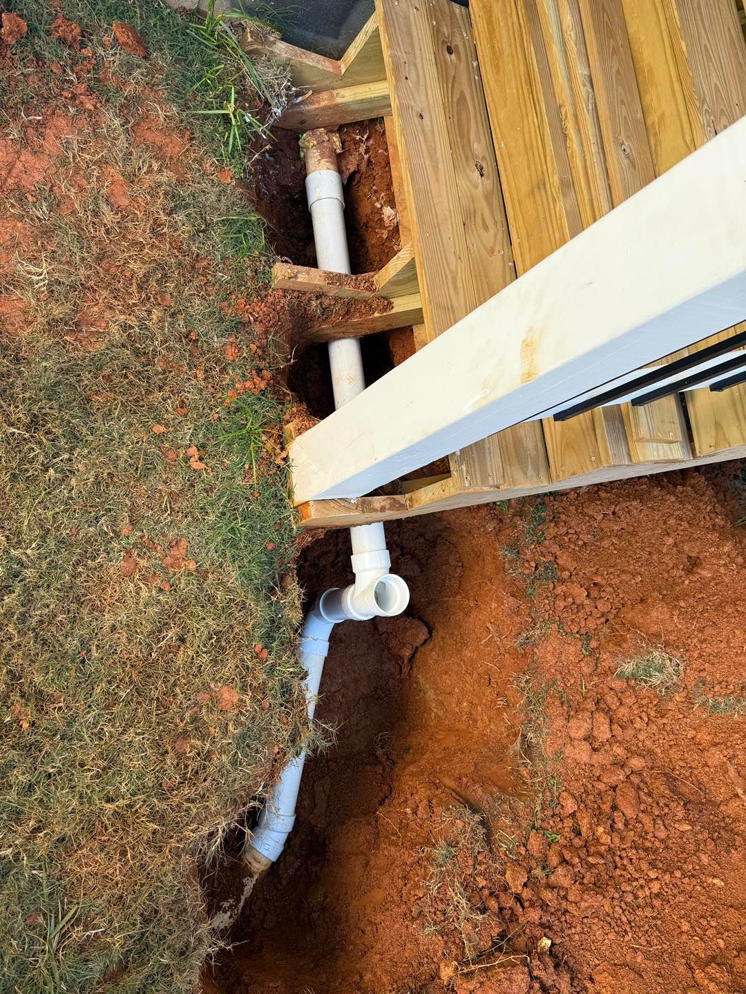 White PVC drainage pipe installed in a trench along the edge of a wooden deck frame and into the yard soil.