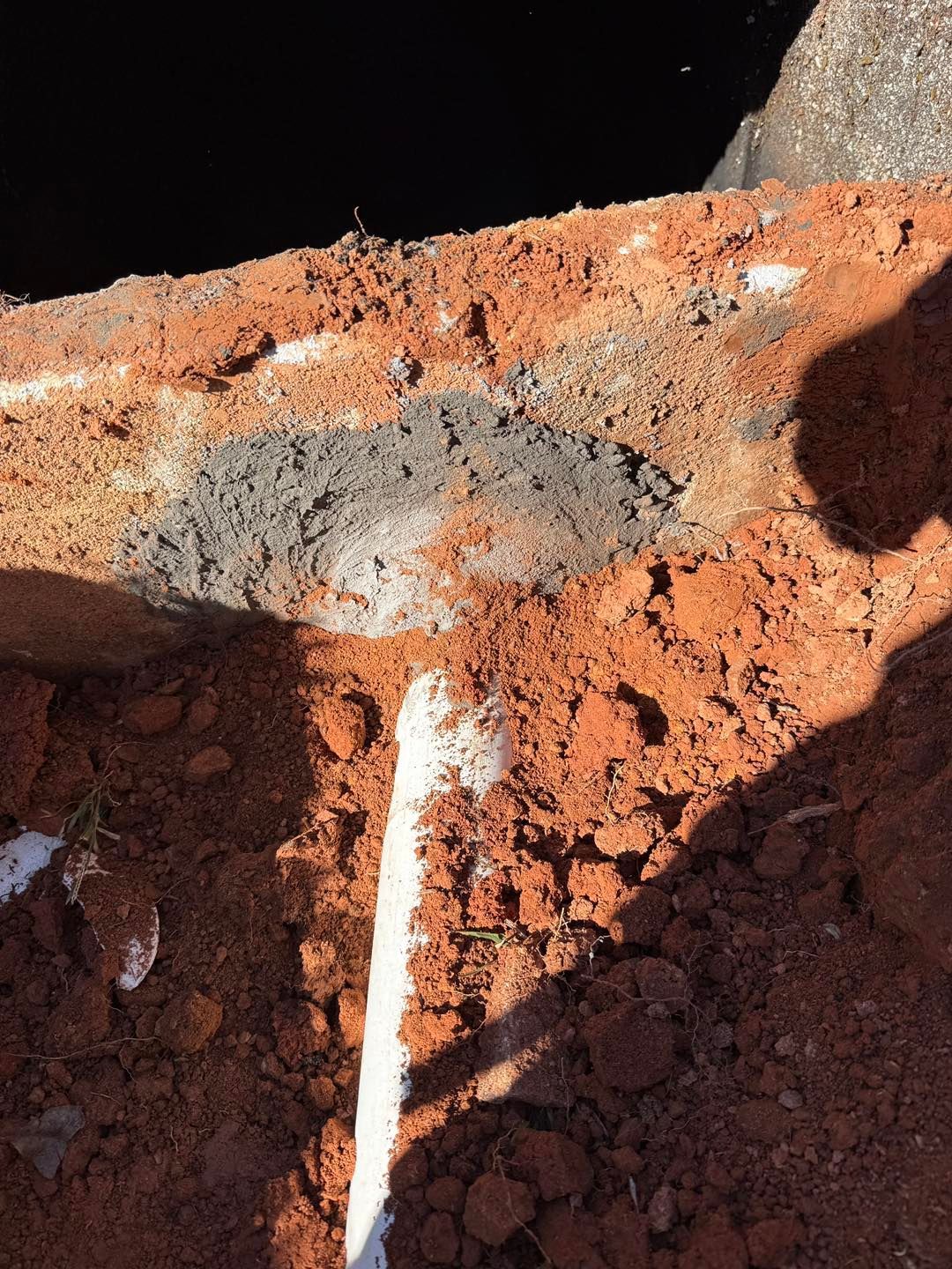 A white PVC pipe enters a hole in a red brick foundation, sealed with grey mortar against a backdrop of dark soil.