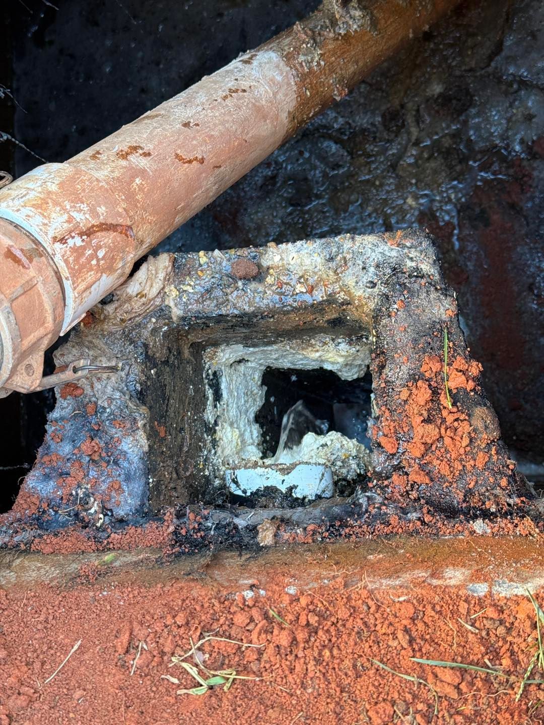 A rectangular concrete drain opening in reddish dirt with a rusted metal pipe positioned above it.