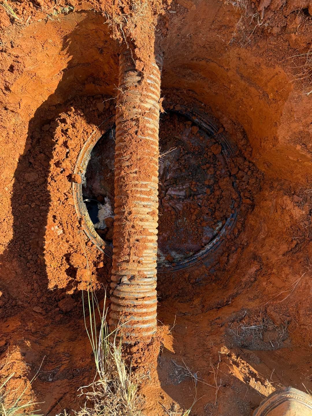 A vertical corrugated metal pipe sits centered in a deep, circular hole in reddish-brown clay soil, partially exposing a lid.