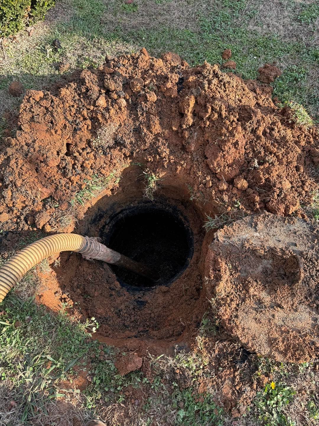 A suction hose inserted into an open, excavated septic tank access port in a grassy yard.