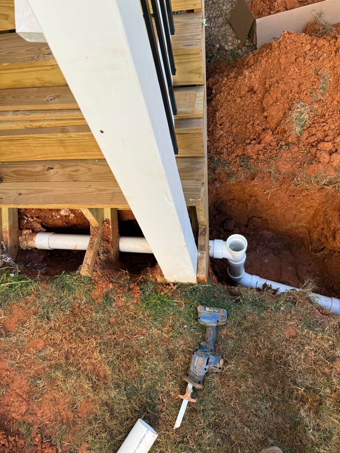 White PVC drainage pipes installed under a wooden deck stairway, with a reciprocating saw lying on the grass nearby.