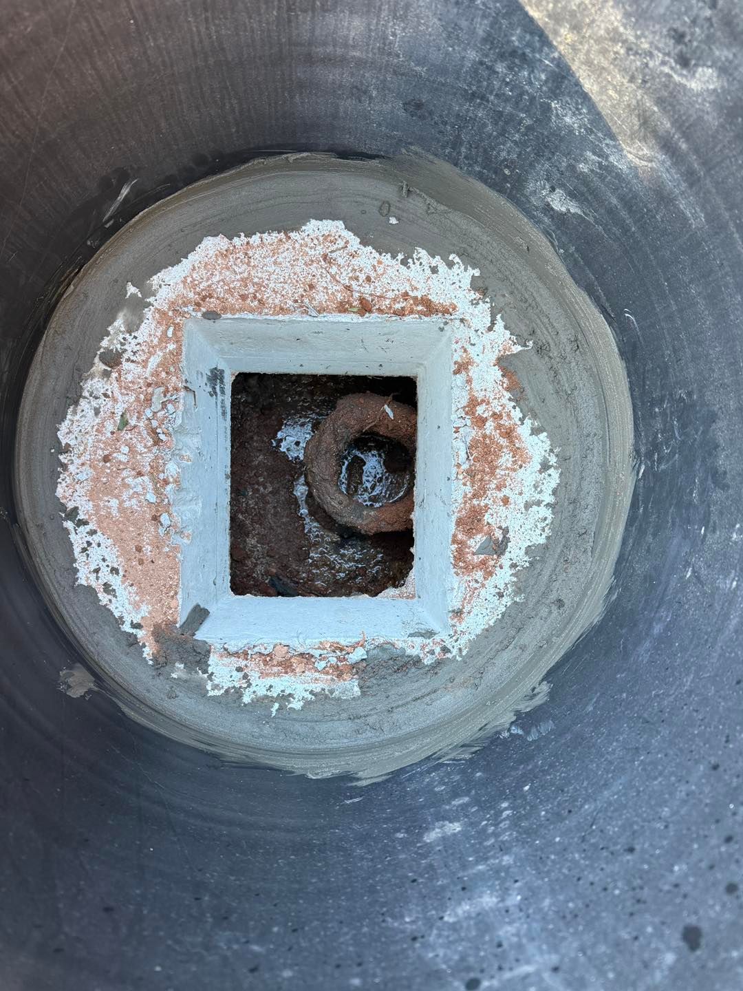 A square drain set in a circular concrete basin with a rusty metal ring visible inside the drain opening.