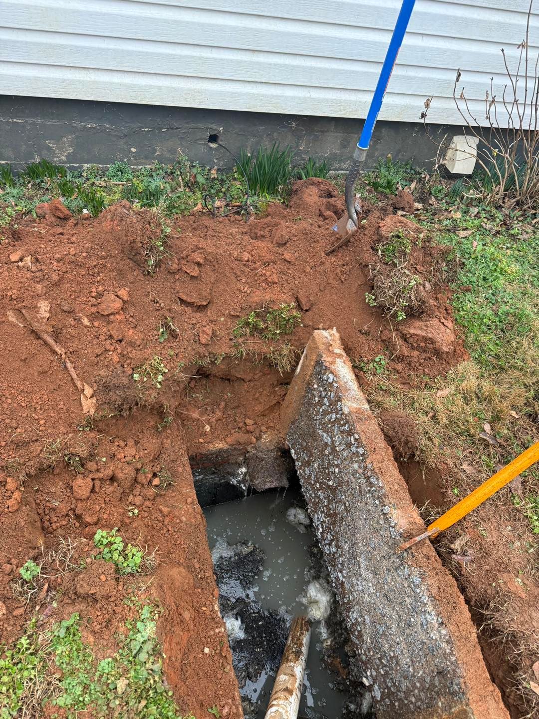 An open, water-filled concrete trench sits in dirt near a house foundation, with two shovel handles visible nearby.