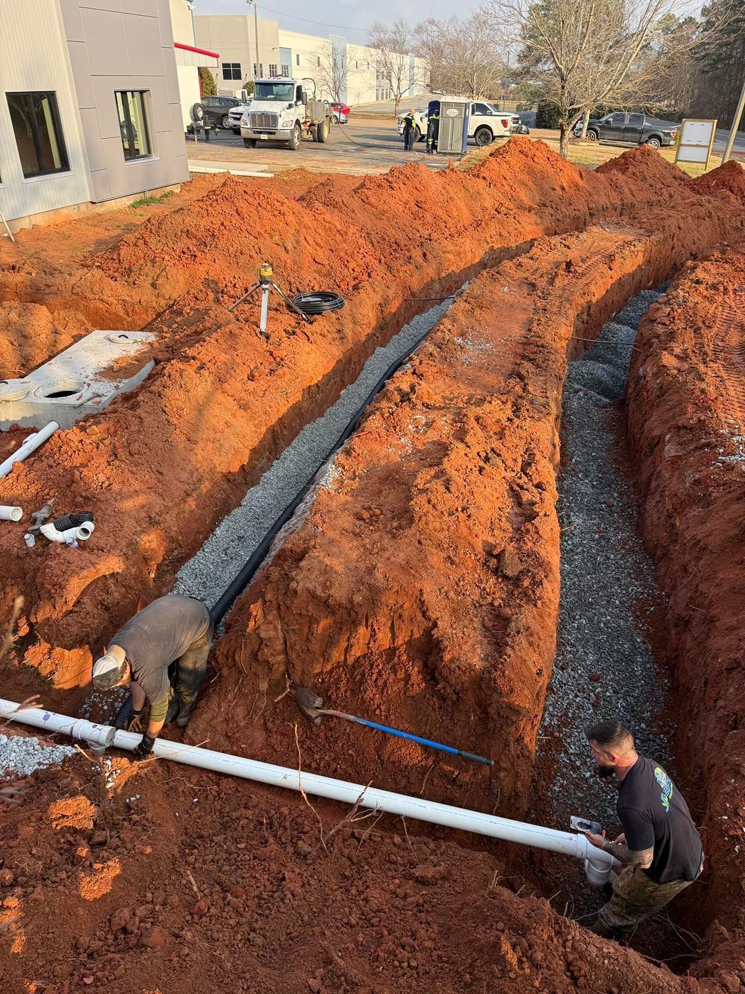 Construction workers installing a drainage system with gravel and white pipes in deep red dirt trenches at a job site.