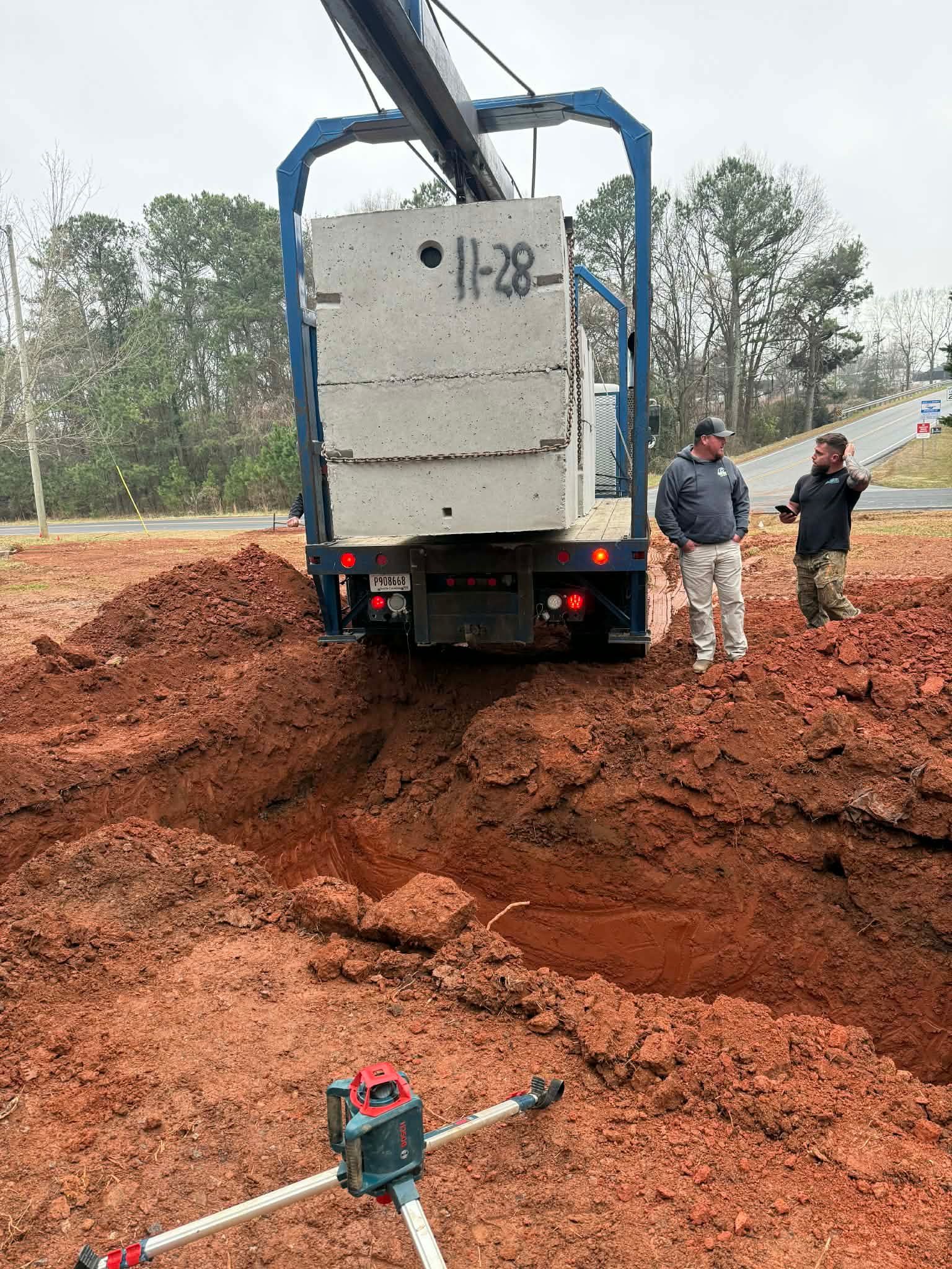 Two construction workers stand by a trench as a crane prepares to lower a large concrete block into an excavated site.