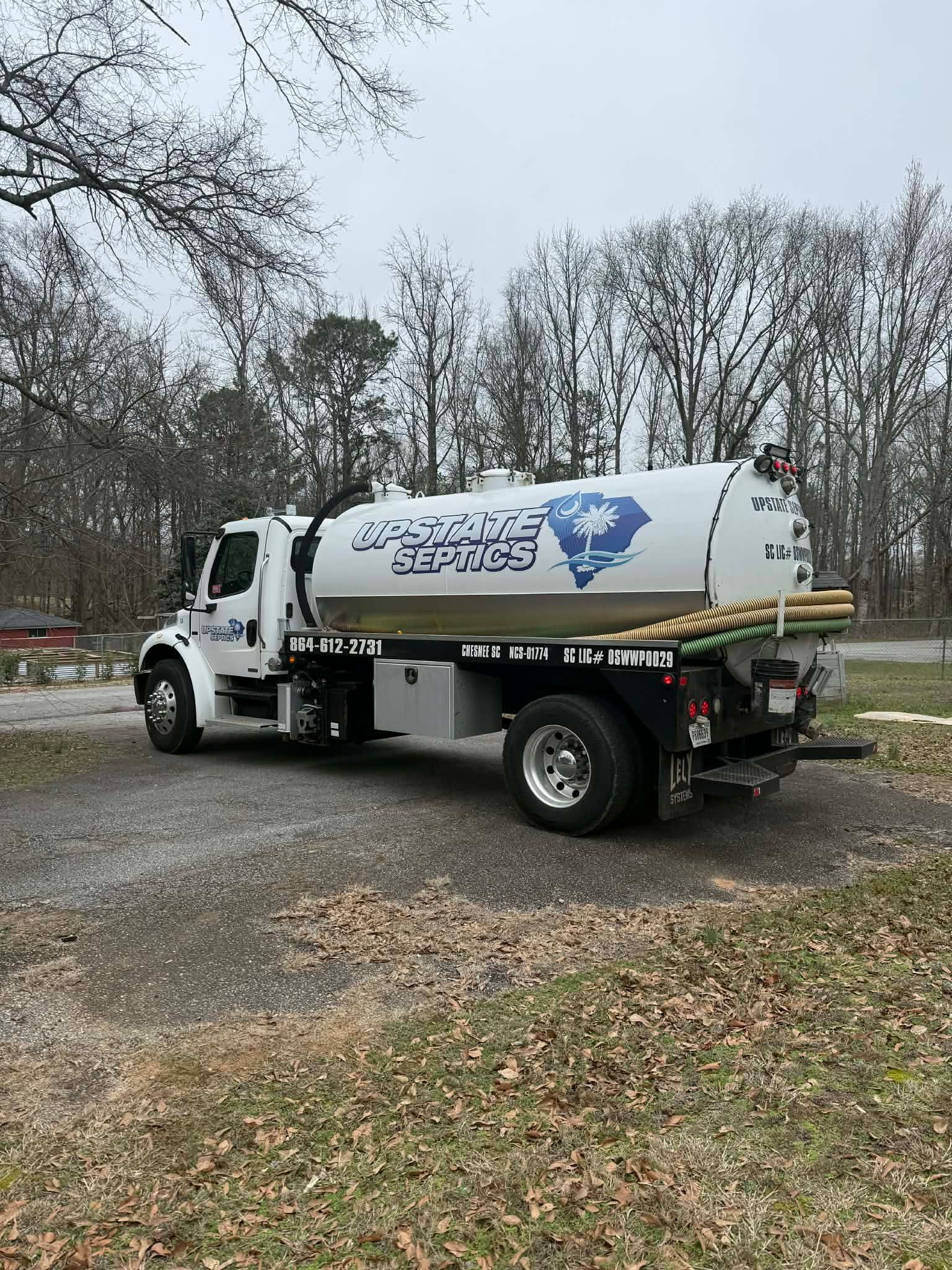A white service truck with a large, cylindrical tank parked on a gravel lot near a wooded area.