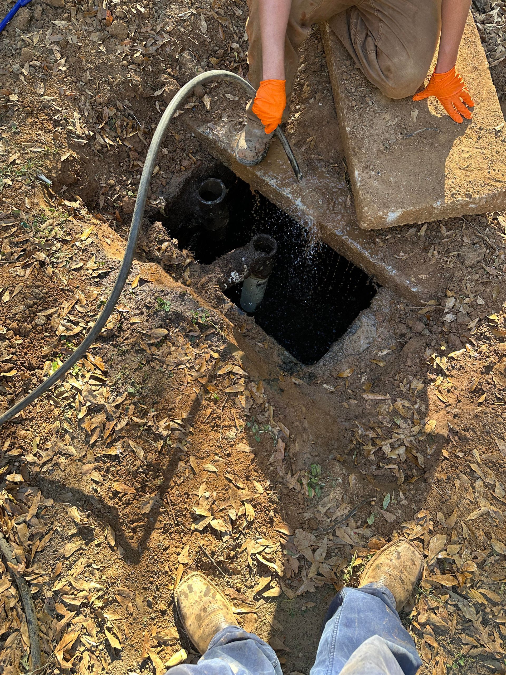 A person in orange gloves uses a hose to spray water into an open, muddy residential septic tank access port.