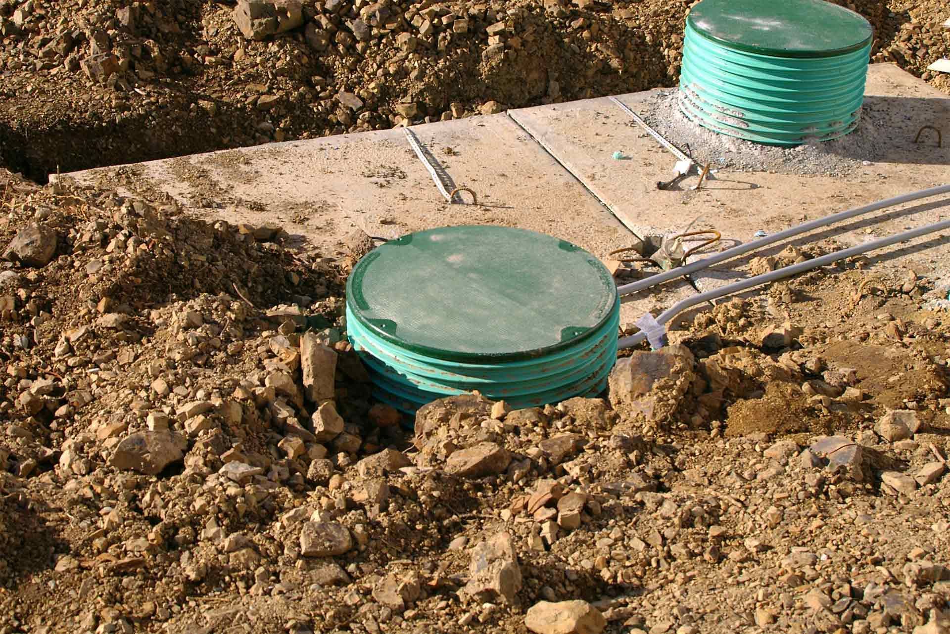 Two green circular access covers for a buried septic system installed on a concrete slab in excavated dirt.