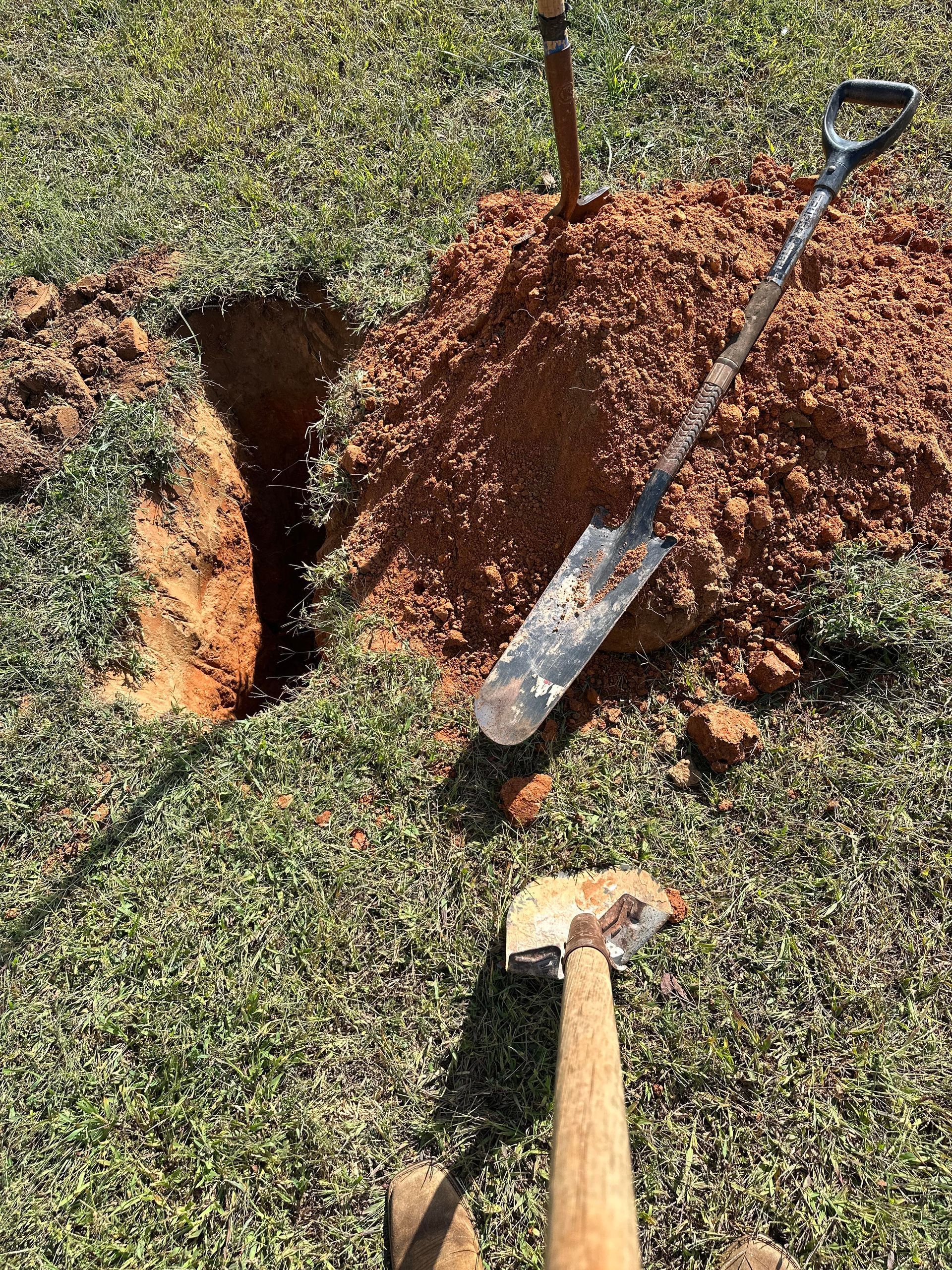 A hole dug in a grassy yard, with a pile of red dirt next to it, an upright shovel, and a second shovel in the foreground.