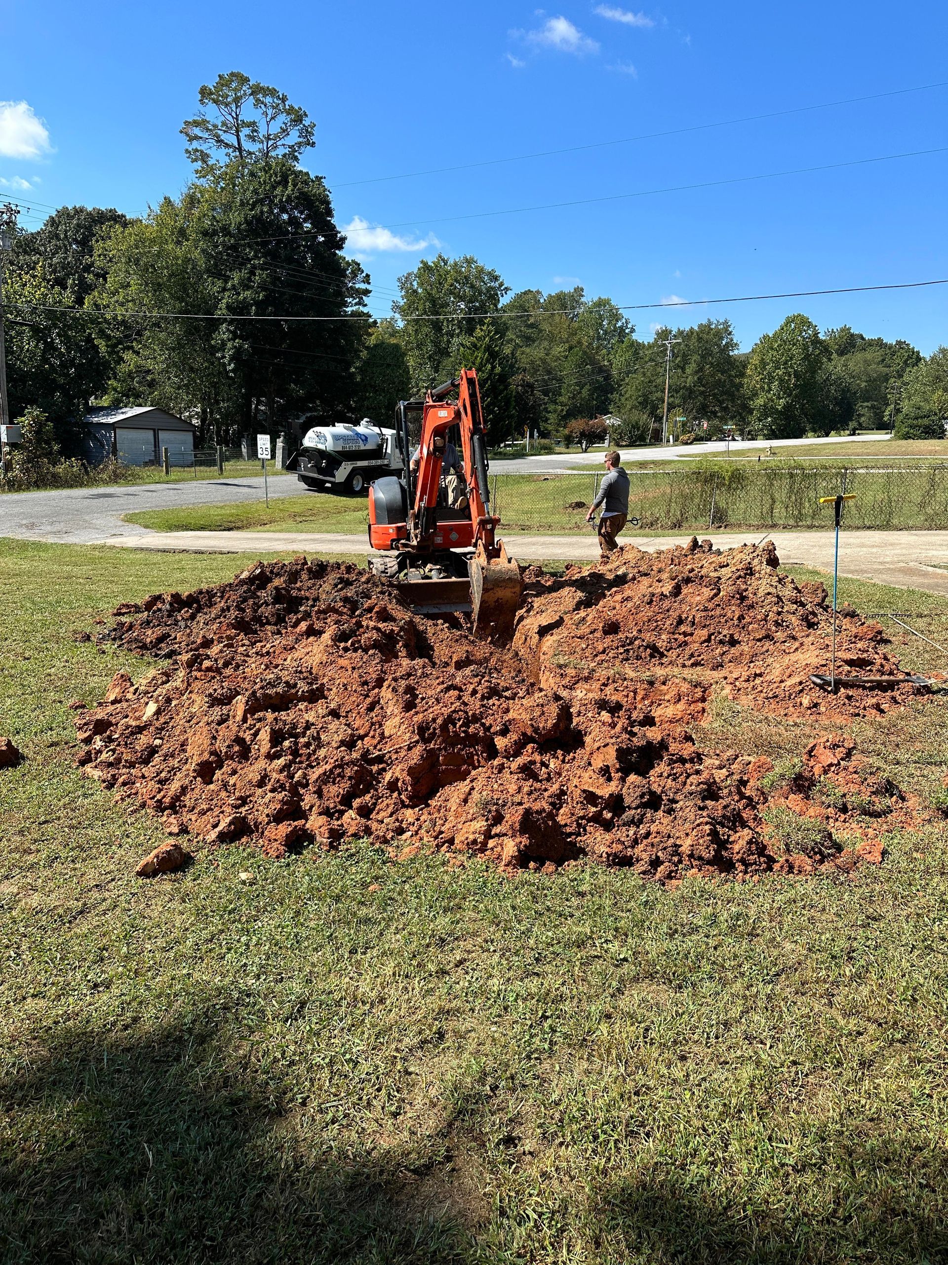 An orange excavator digs in a dirt pile on a grassy lot under a bright blue sky.