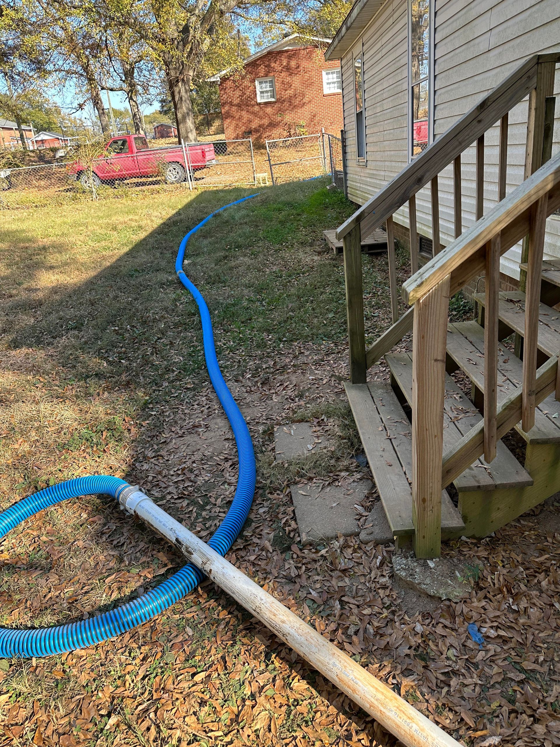 A long blue hose snakes across a leaf-covered lawn toward the wooden stairs of a house, with a white pipe in the foreground.