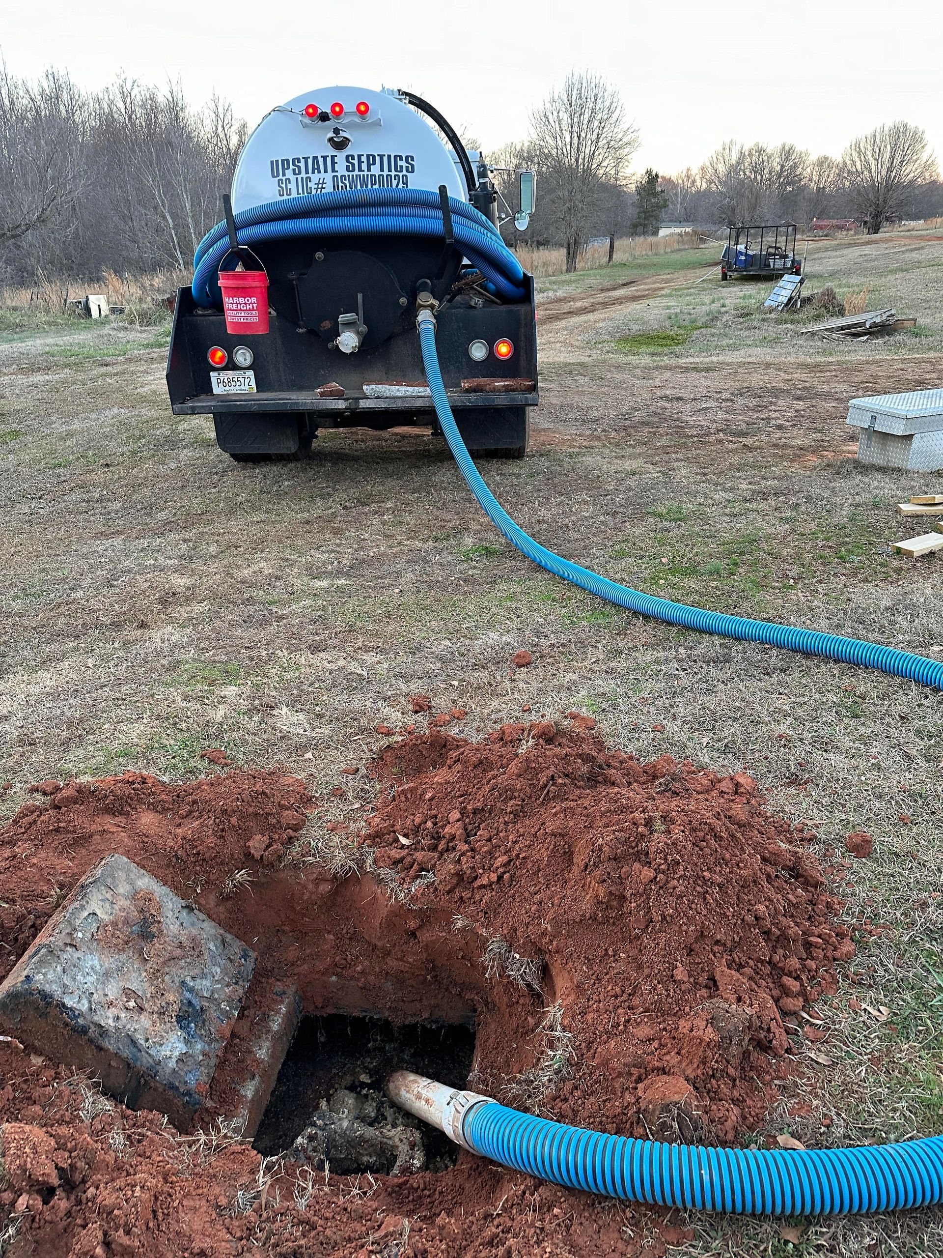 A septic service truck connected by a blue hose to an open, excavated septic tank in a rural field.