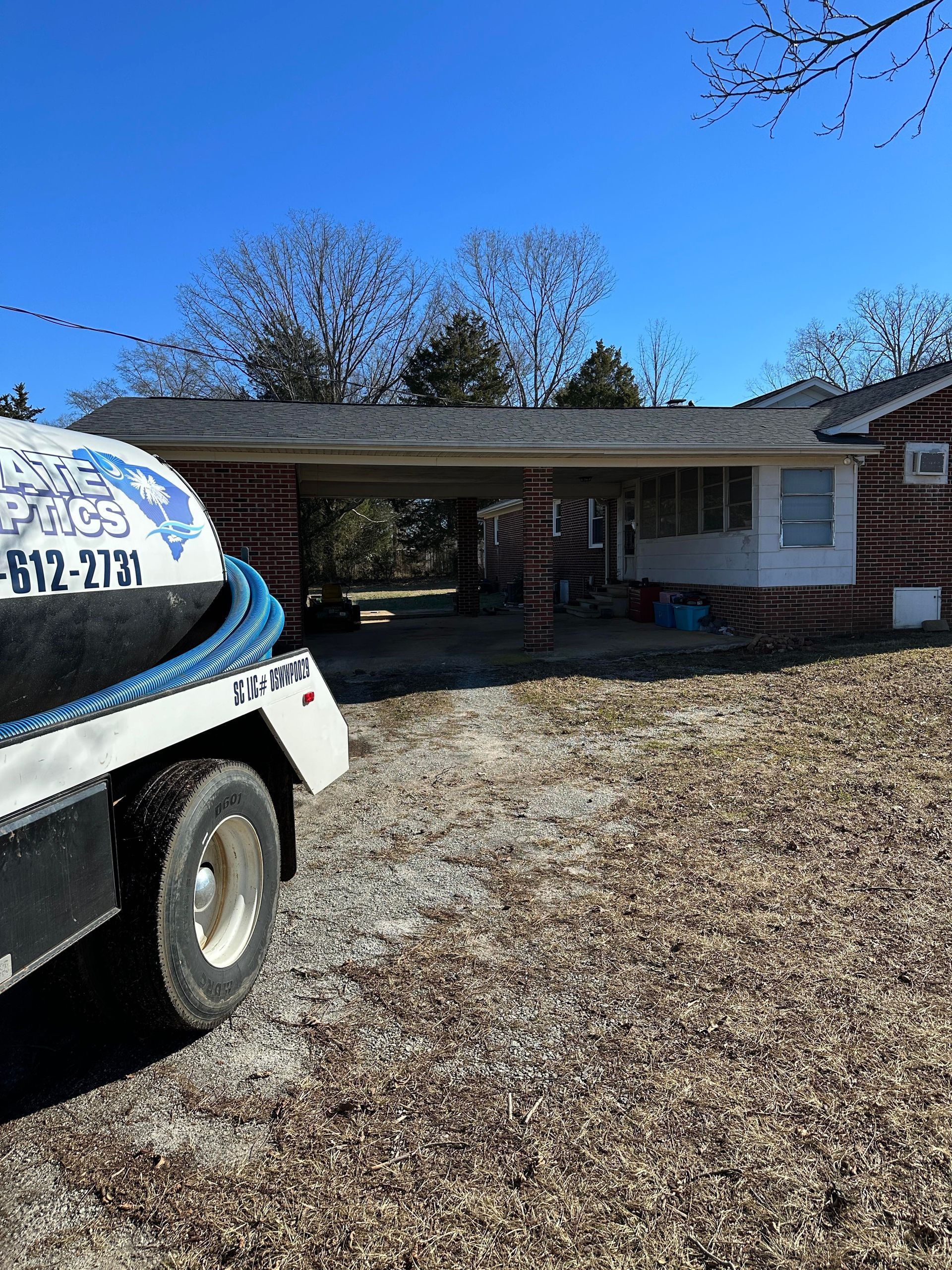 A large, white service truck with a hose parked on a gravel driveway in front of a house with a carport.