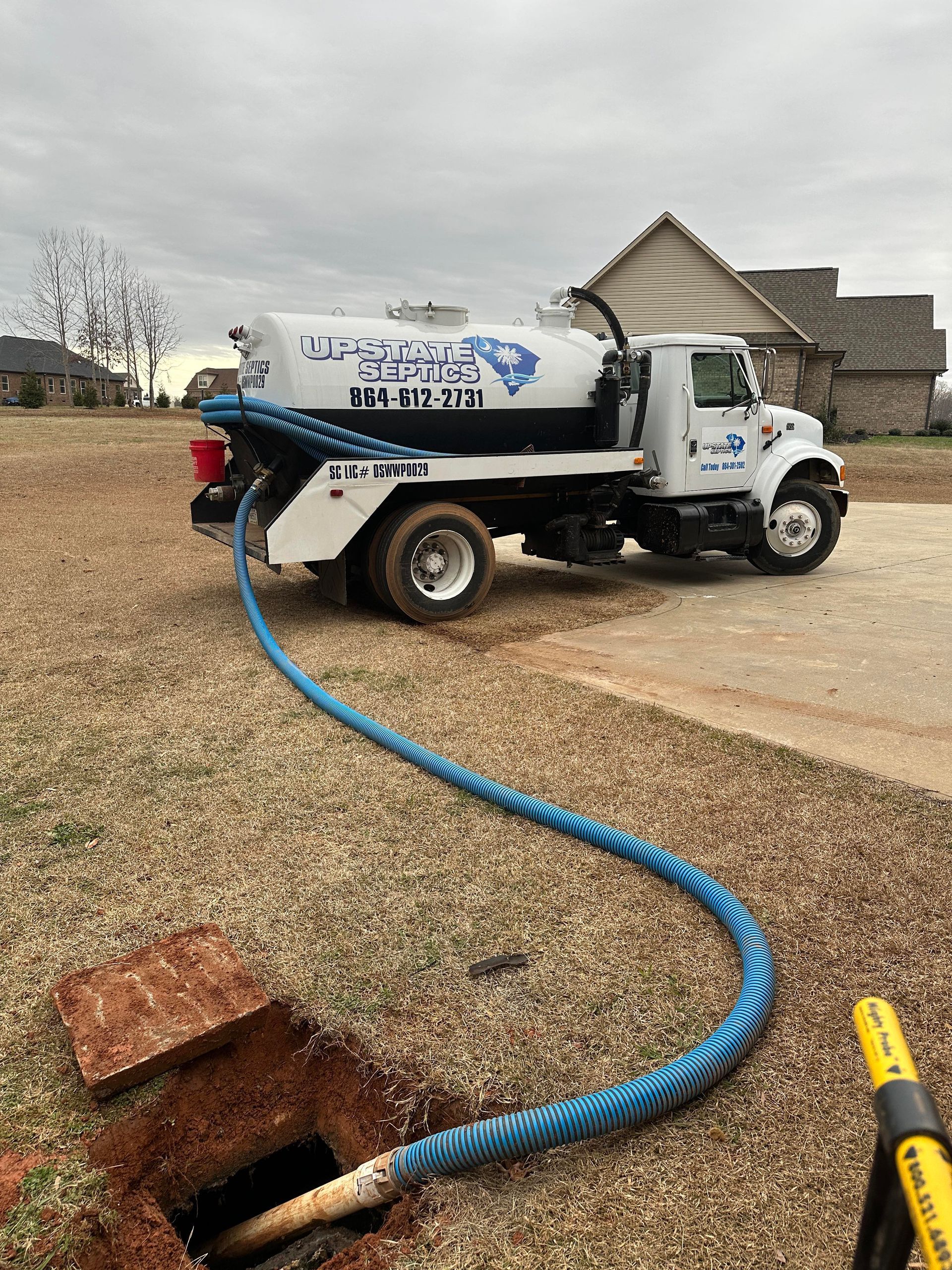 A white septic truck parked on a gravel lot with a blue hose running into an open underground septic tank.