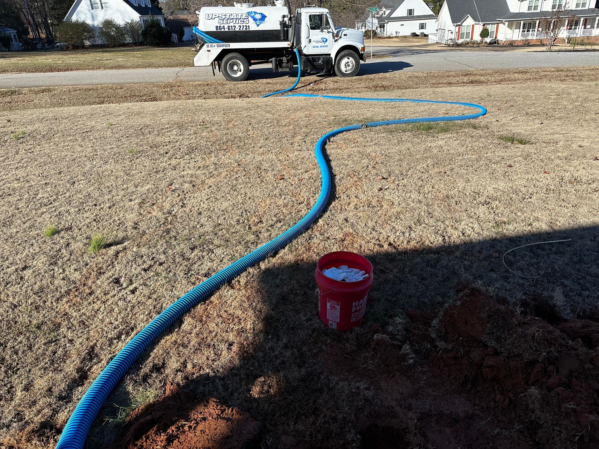 A white tanker truck parked on a street with a long blue hose extending across a dry lawn to a red bucket.