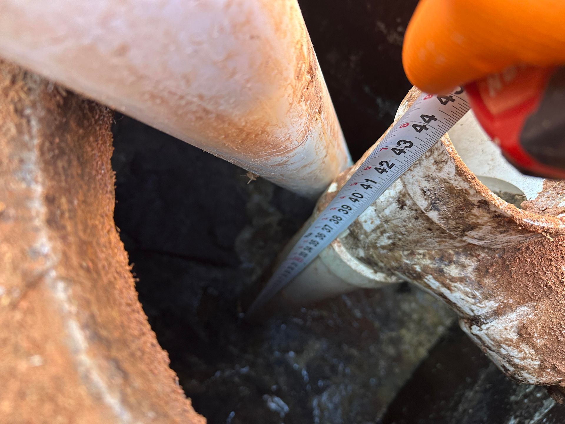 A gloved hand uses a tape measure to gauge the depth of a pipe inside a dark, open concrete access hole.