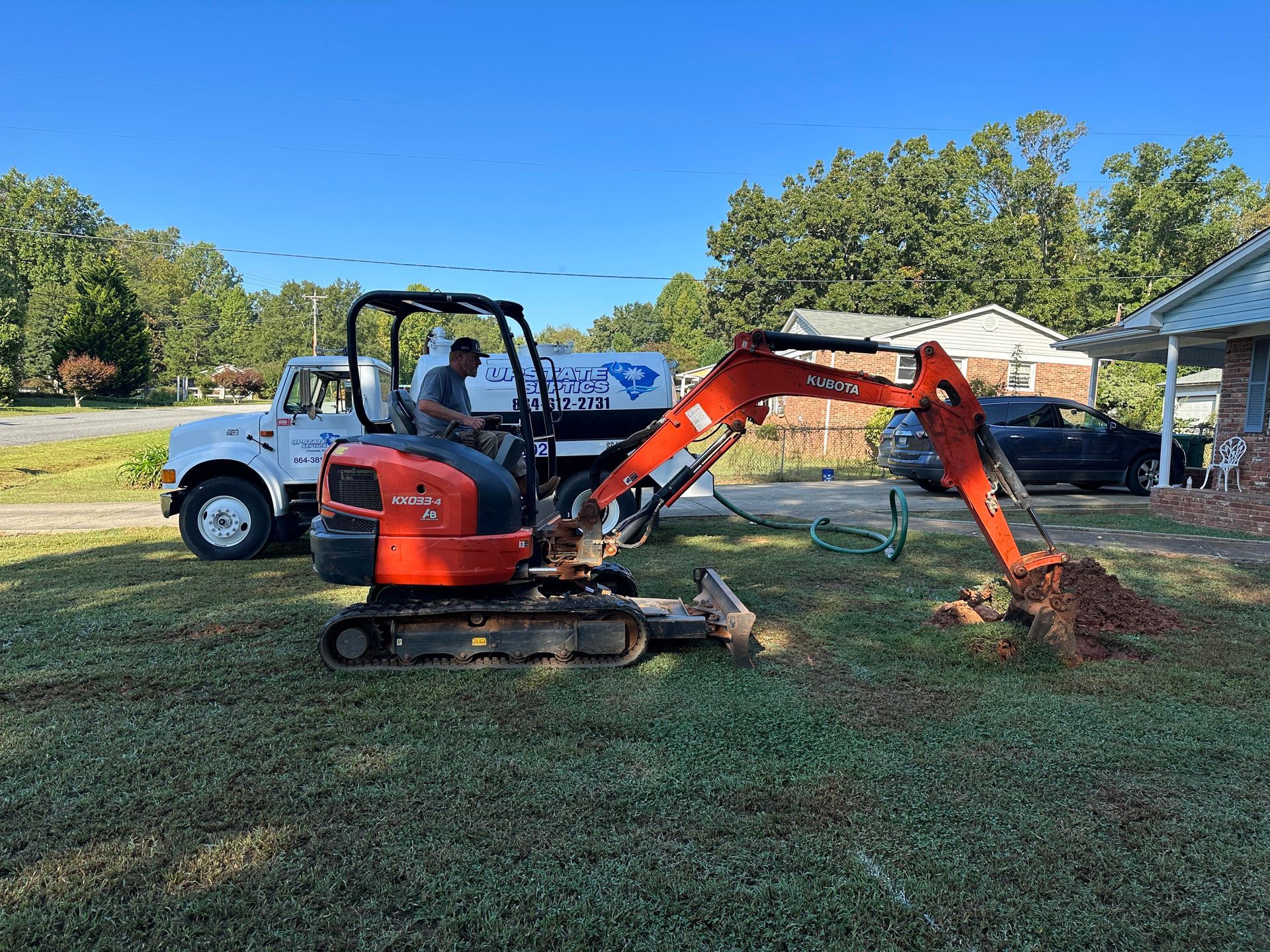 An orange mini excavator digging in a residential yard with a utility truck and house in the background.