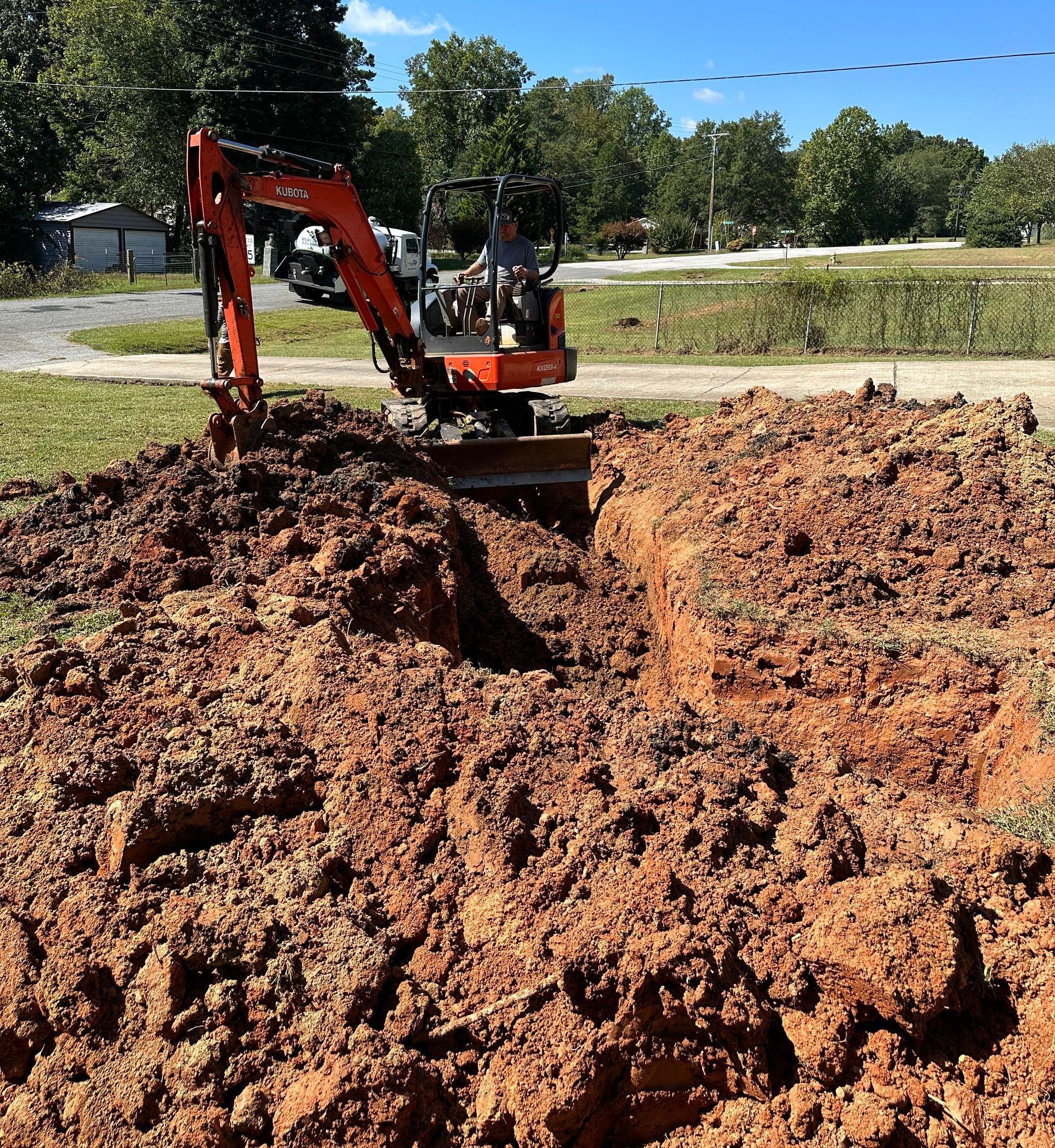 An orange mini-excavator digs a deep trench in reddish-brown soil on a bright, sunny day.