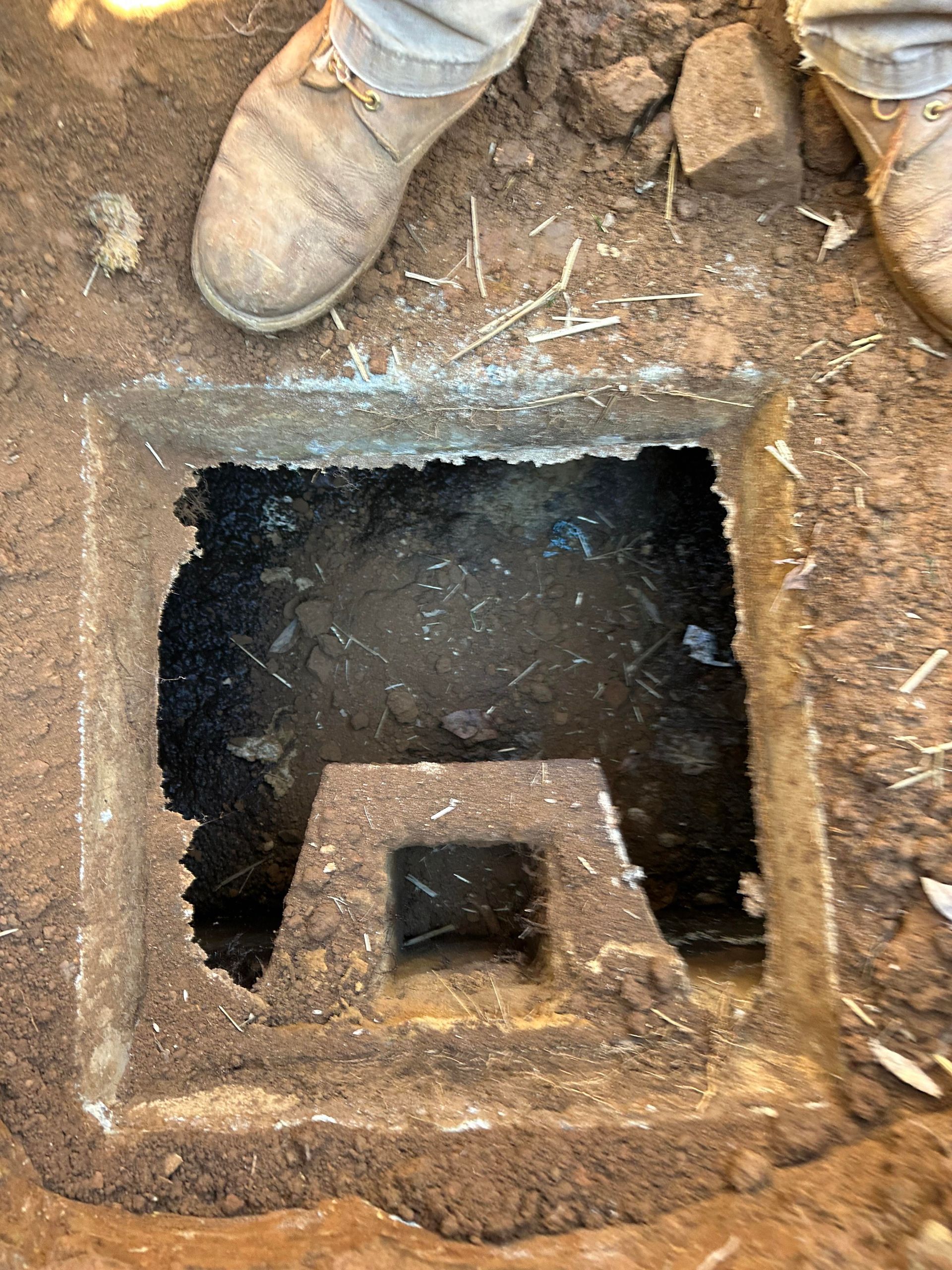 Top-down view of a rectangular, open concrete drainage box embedded in dirt, with muddy water at the bottom.
