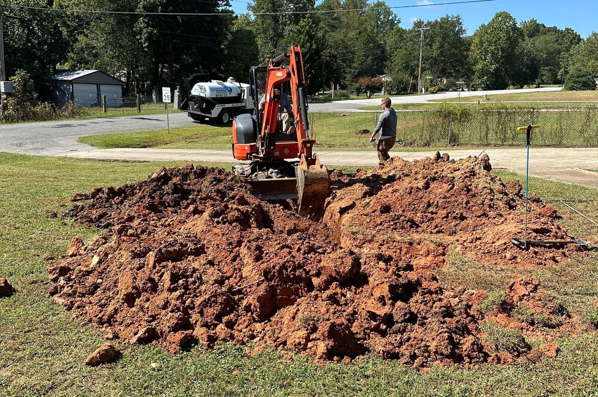 An orange excavator digs a trench in a grassy, dirt-filled field with a person standing nearby.
