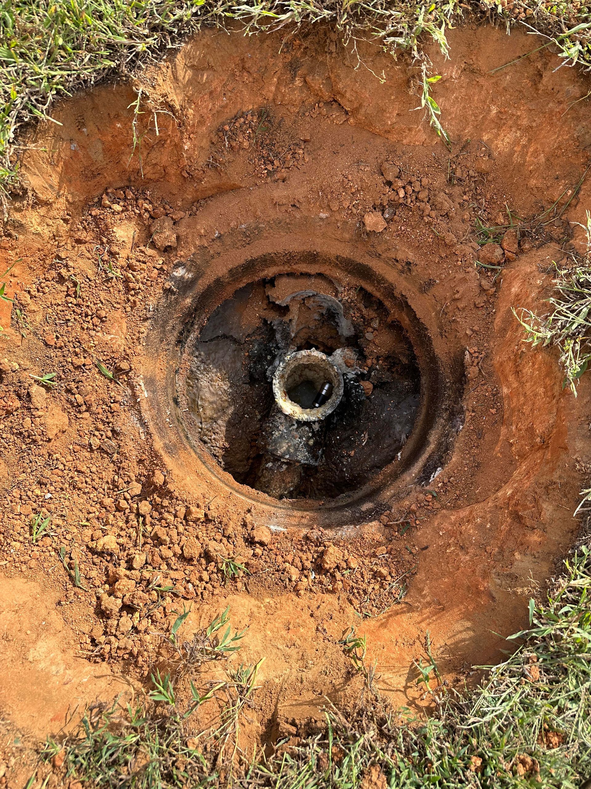 An open circular utility access hole in reddish-brown dirt, revealing an internal pipe and dark, muddy debris.