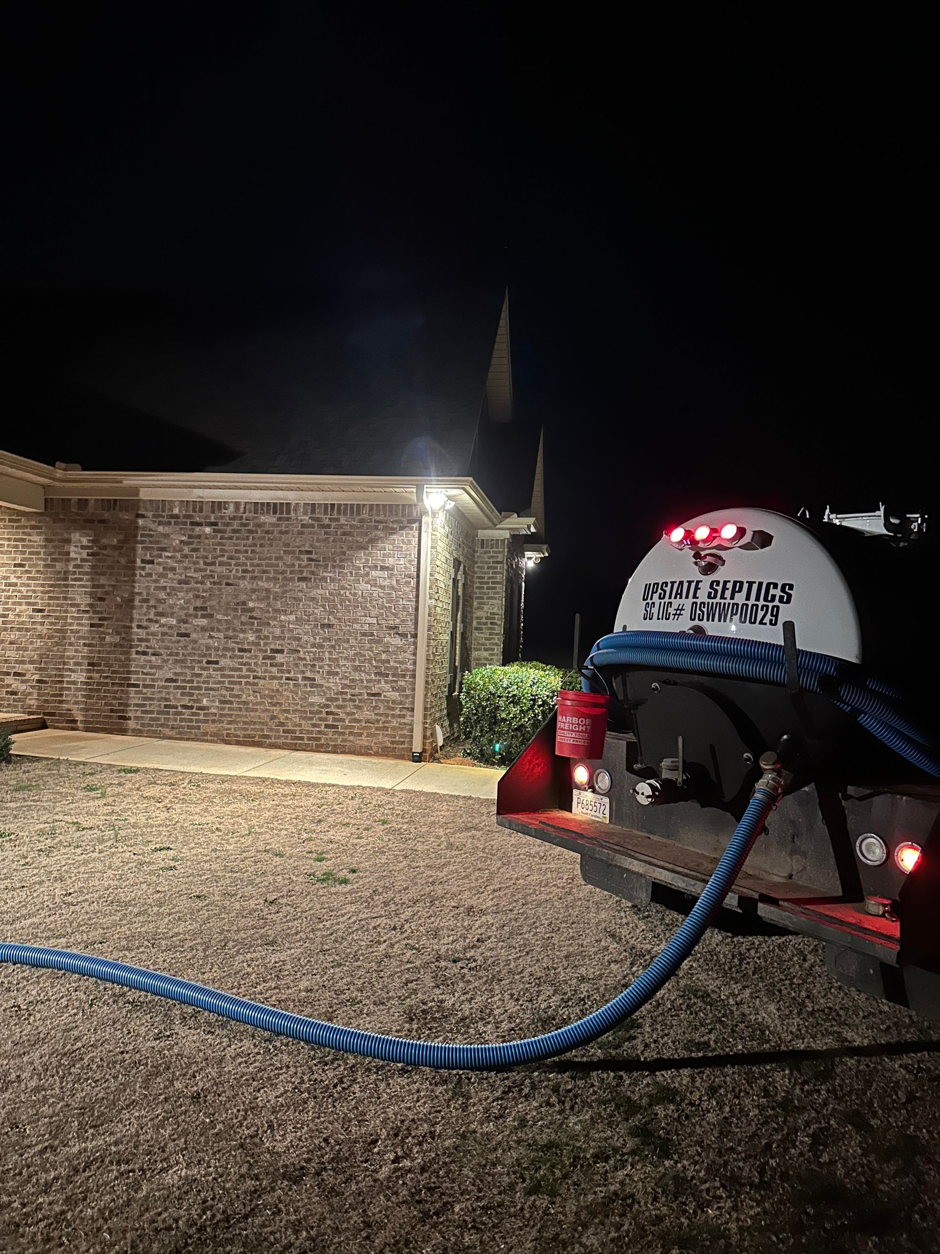 A trailer with a white tank and a blue hose sits at night on a gravel driveway beside a lit brick house.