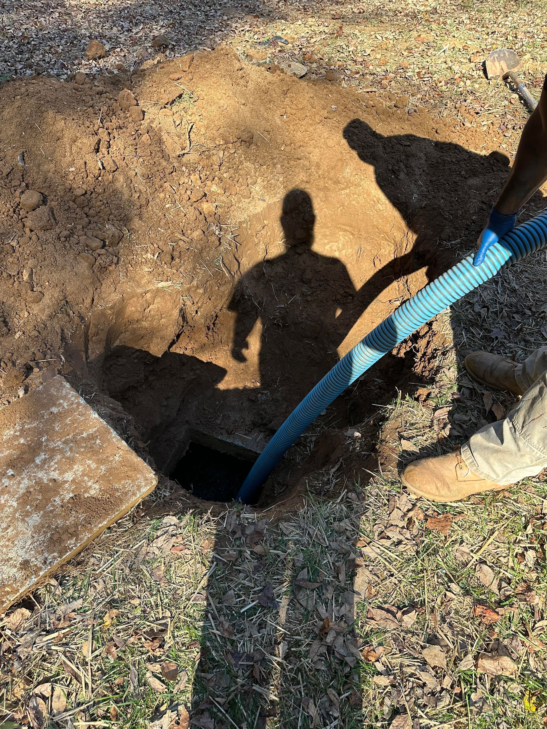 A person in work boots uses a blue hose to pump out a square, dark hole in the ground covered by a concrete slab.