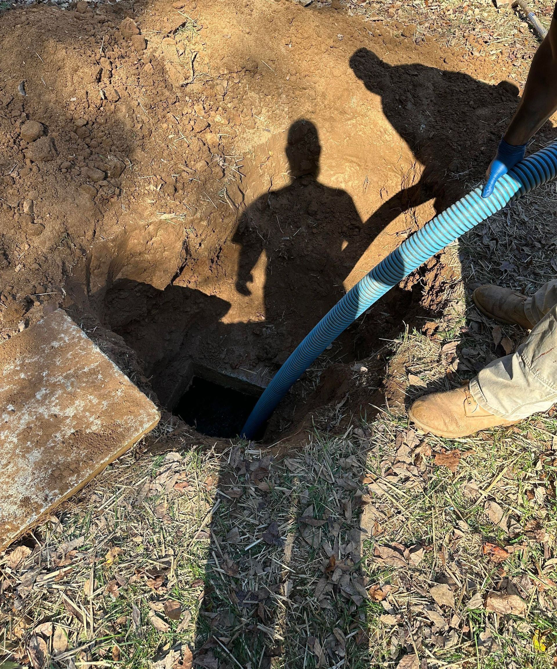 A worker uses a blue hose to vacuum out a hole in the ground, with their shadow cast over the excavated area.