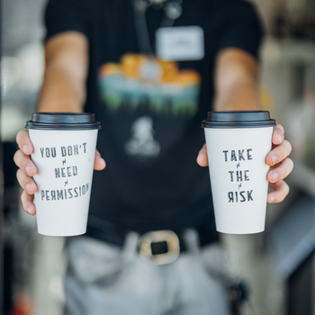 Two takeaway cups held out, one reading “You don’t need permission” and the other “Take the risk.”