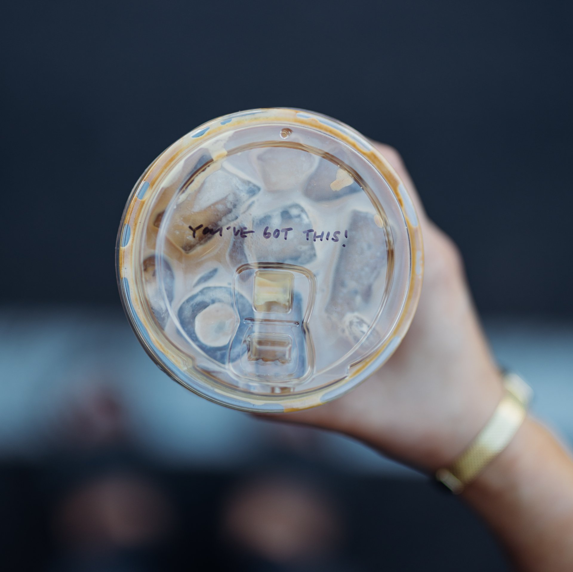 Reusable cup lid held up against a blurred dark background, with “you me got this” written on it