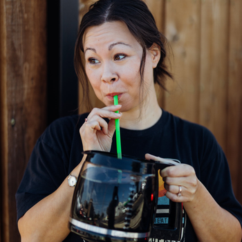 Woman sipping from a large black mug with a green straw, standing indoors by a wooden wall