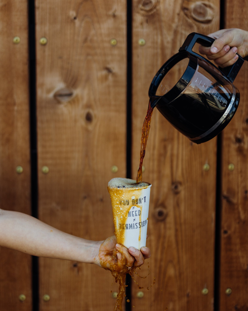 Coffee being poured into a clear plastic cup held by a hand, against a wooden fence background.
