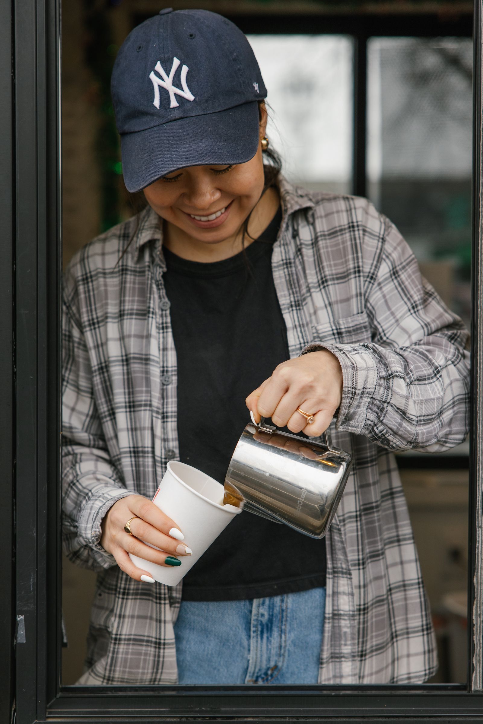 Person in a navy cap pours coffee into a white cup by a window.