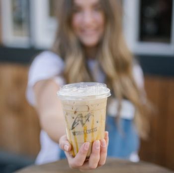Person holding out an iced coffee in a clear plastic cup with a straw indoors.
