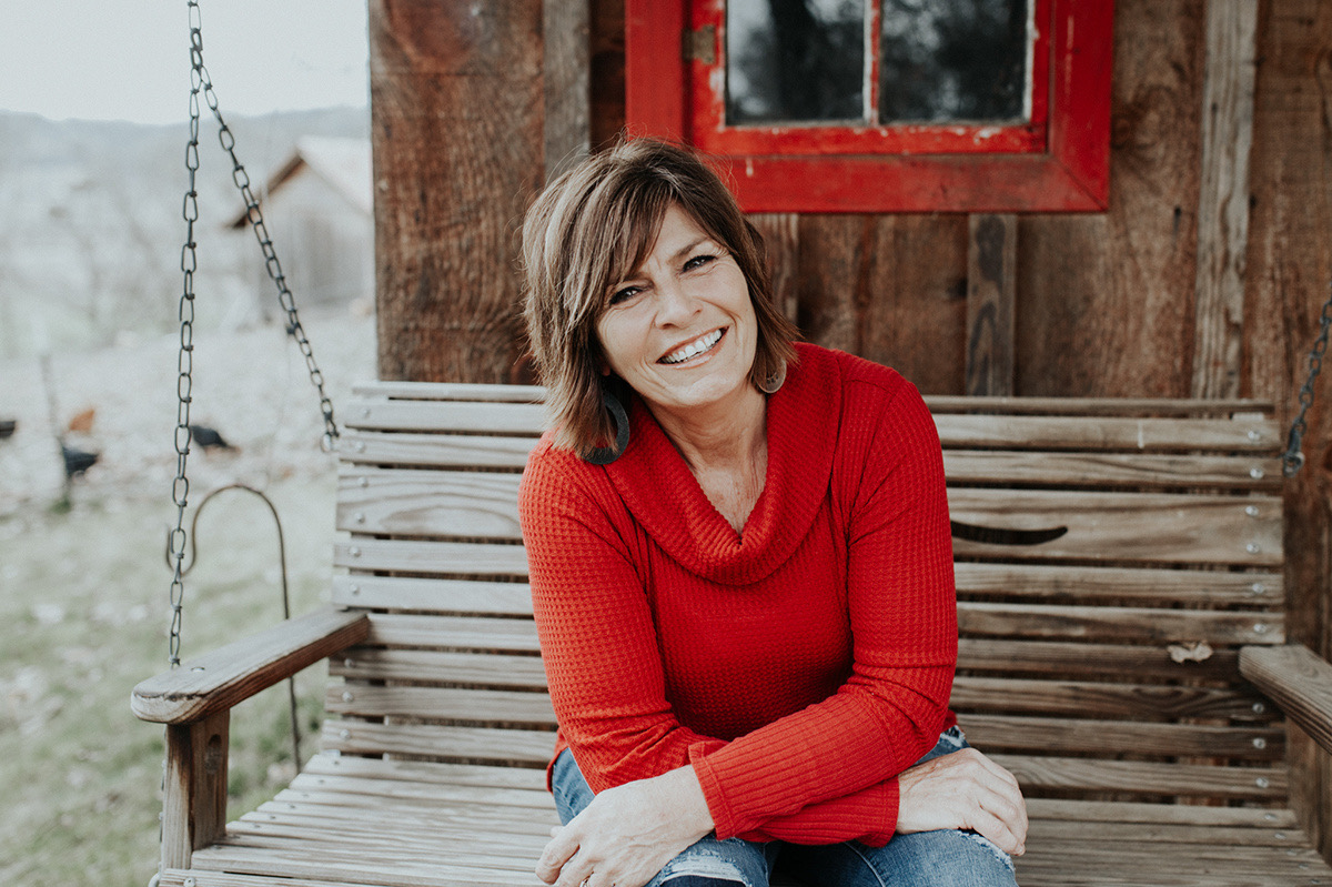 Smiling woman in a red sweater sitting on a wooden porch swing in front of a rustic cabin