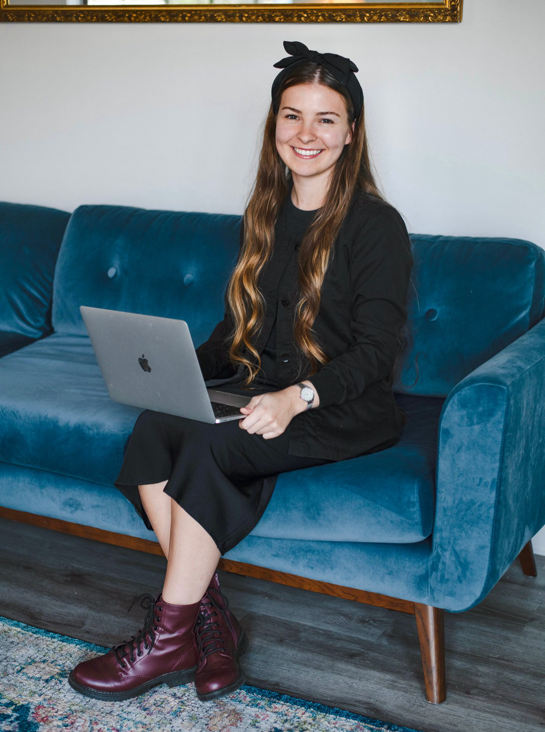 Woman sitting on a teal sofa with a laptop, wearing a black outfit and burgundy boots, smiling at the camera