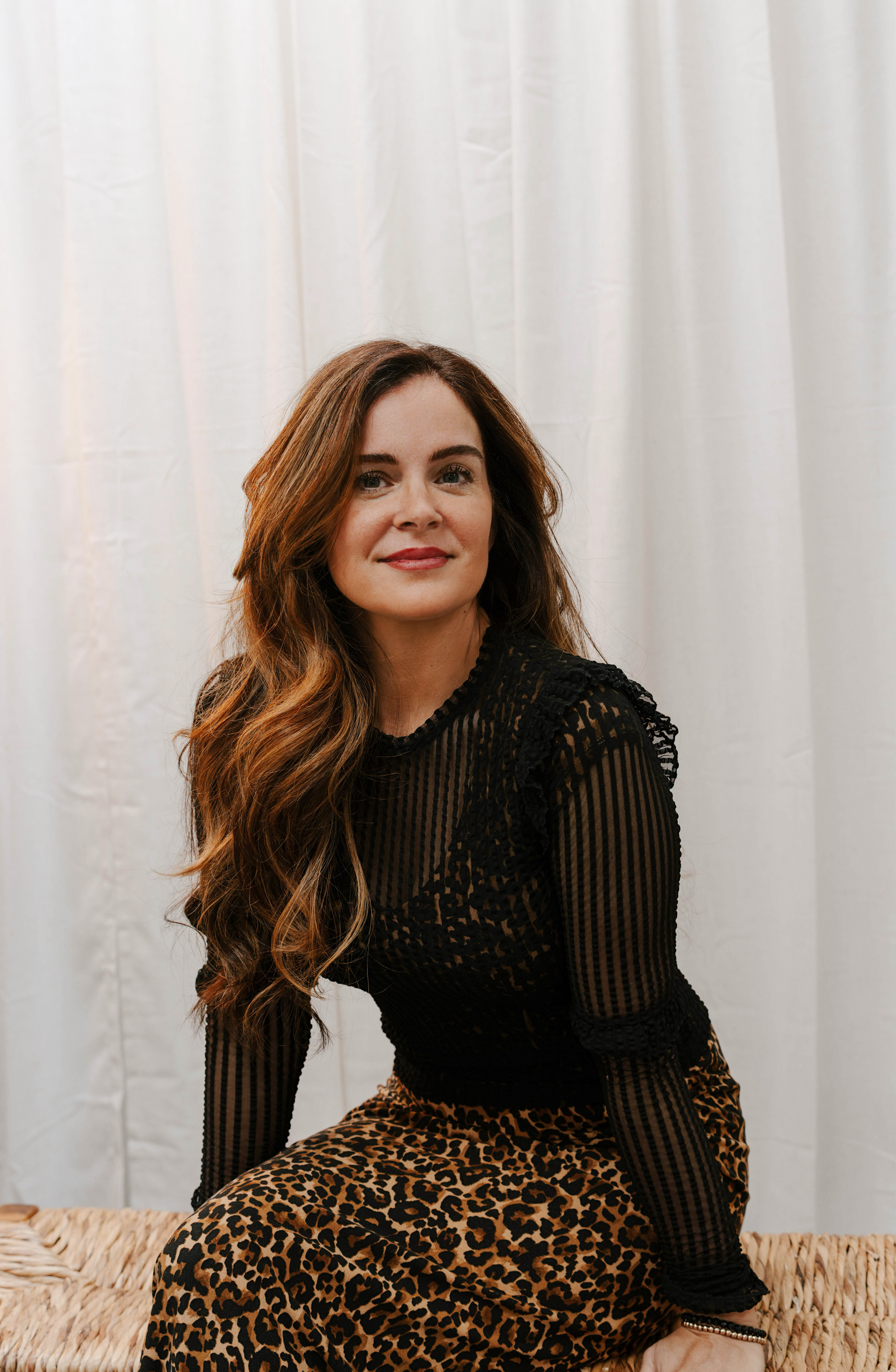 Woman in a black sheer top and leopard skirt seated against a white curtain backdrop.