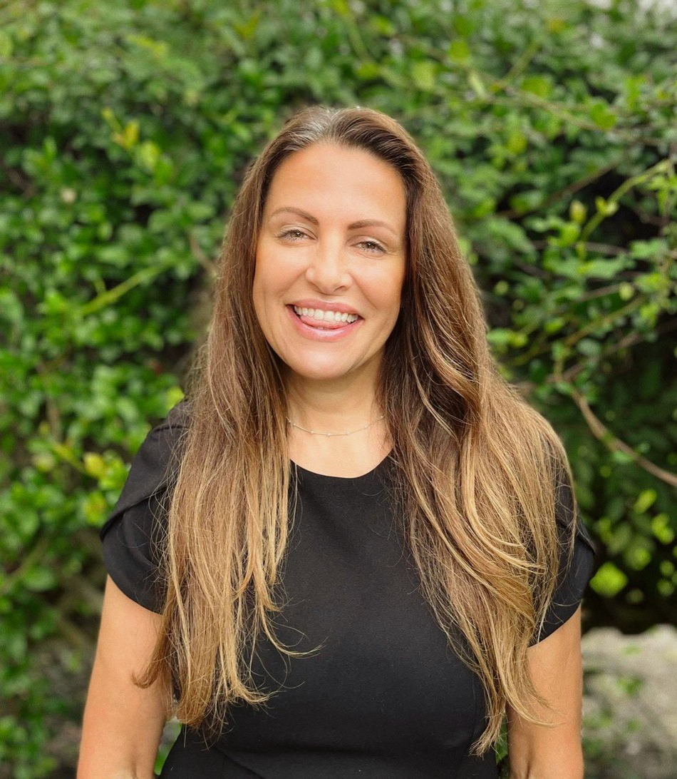 Smiling woman with long hair in a black top standing in front of green bushes