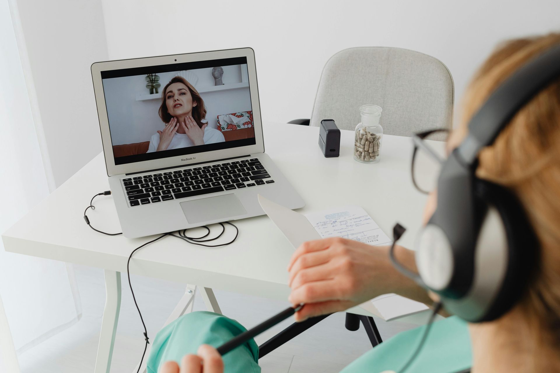 Person wearing headphones on a video call, seated at a white desk with a laptop and notebooks.