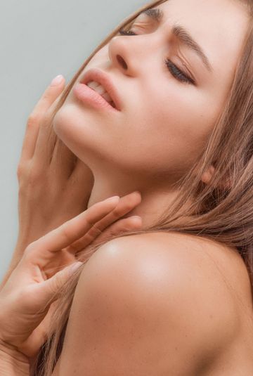 Close-up of a woman with closed eyes touching her face and shoulder against a soft gray background