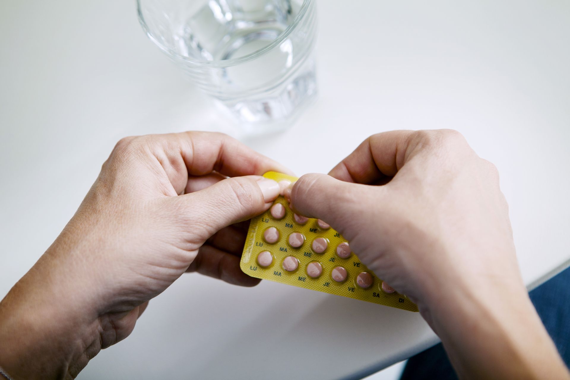Hands opening a yellow blister pack of pills beside a glass of water on a white table