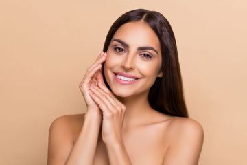 Smiling woman with long dark hair, resting her hand on her cheek against a beige background