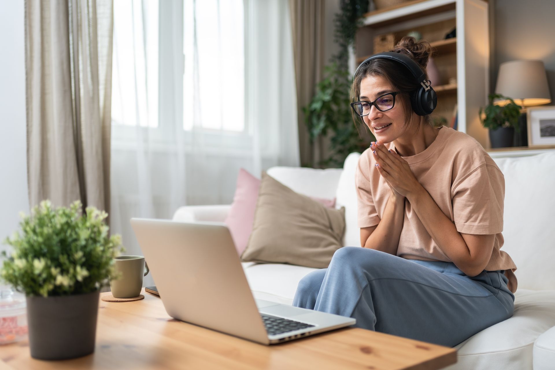 Person with headphones smiling on a couch, using a laptop in a bright living room.