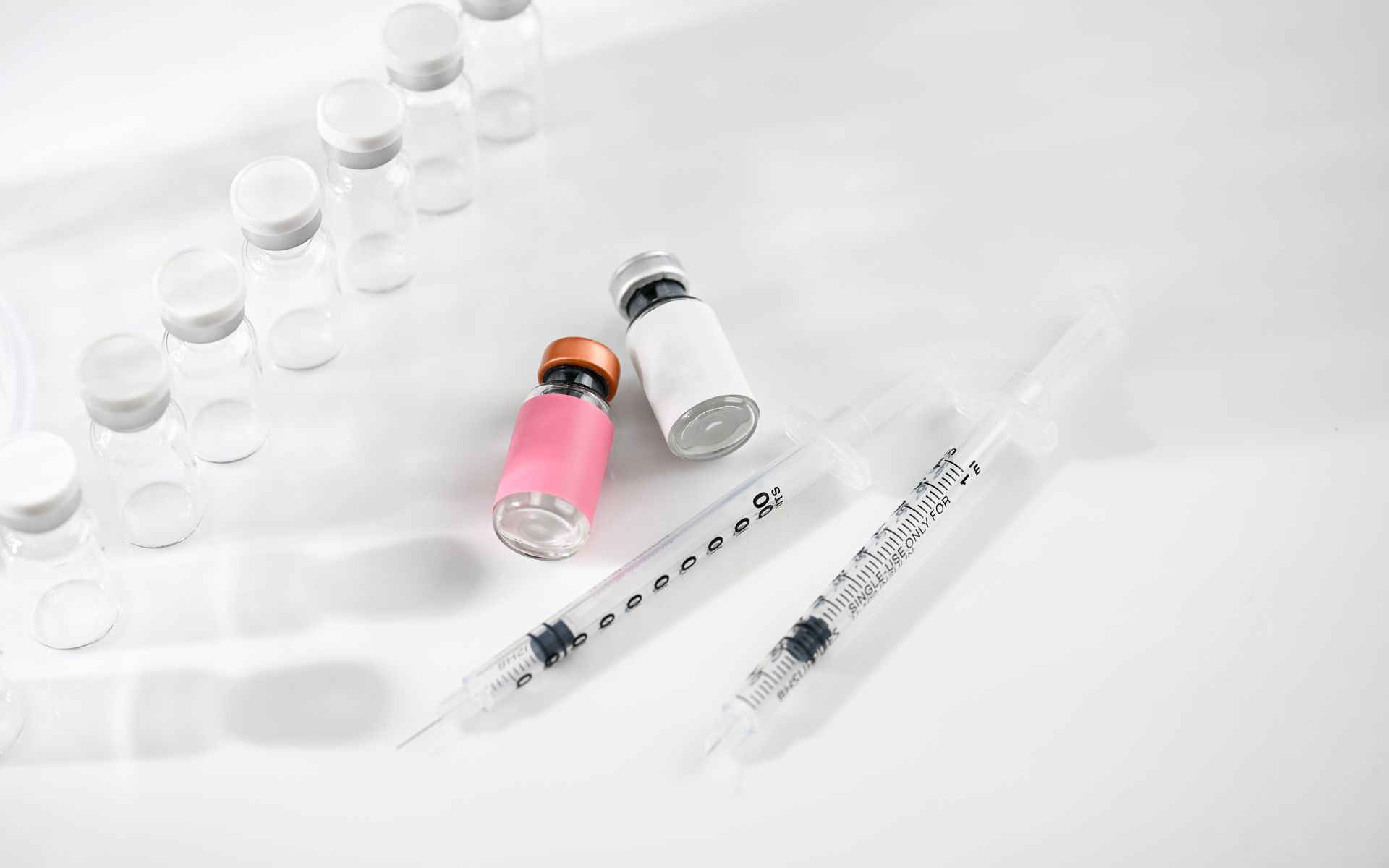 Assorted vaccine vials and syringes on a white surface, including pink and clear vials.