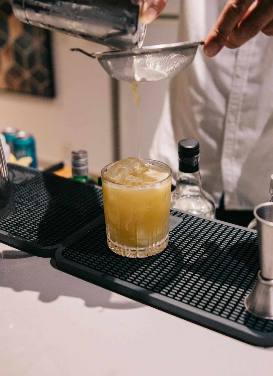 Cocktail being strained into a glass on a bar counter, with bottles and bar tools nearby