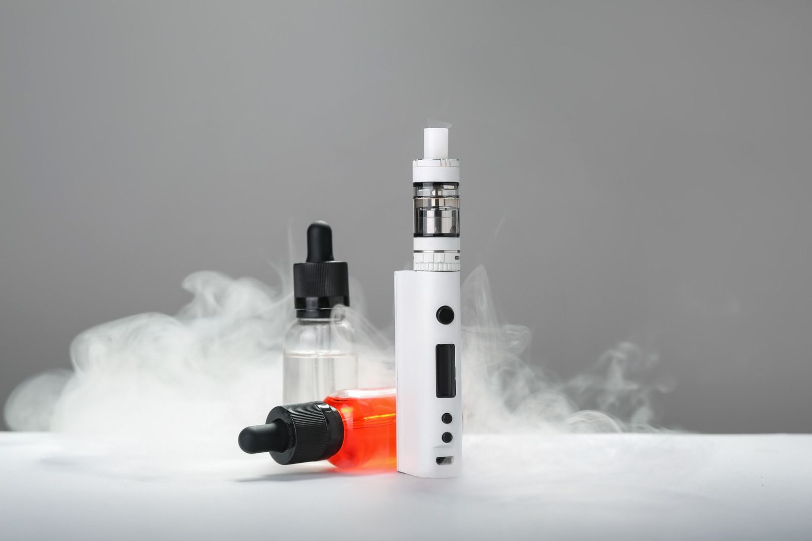 Vape devices and e-liquid bottles surrounded by white vapor on a gray background