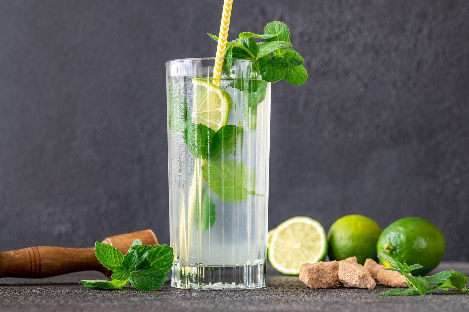 Tall glass of mojito with lime, mint, and straw on a dark table with whole limes and ginger root